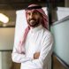 Middle Eastern male professional in traditional clothing standing in meeting room. Happy young Saudi businessman standing with arms crossed looking at camera and smiling in office.